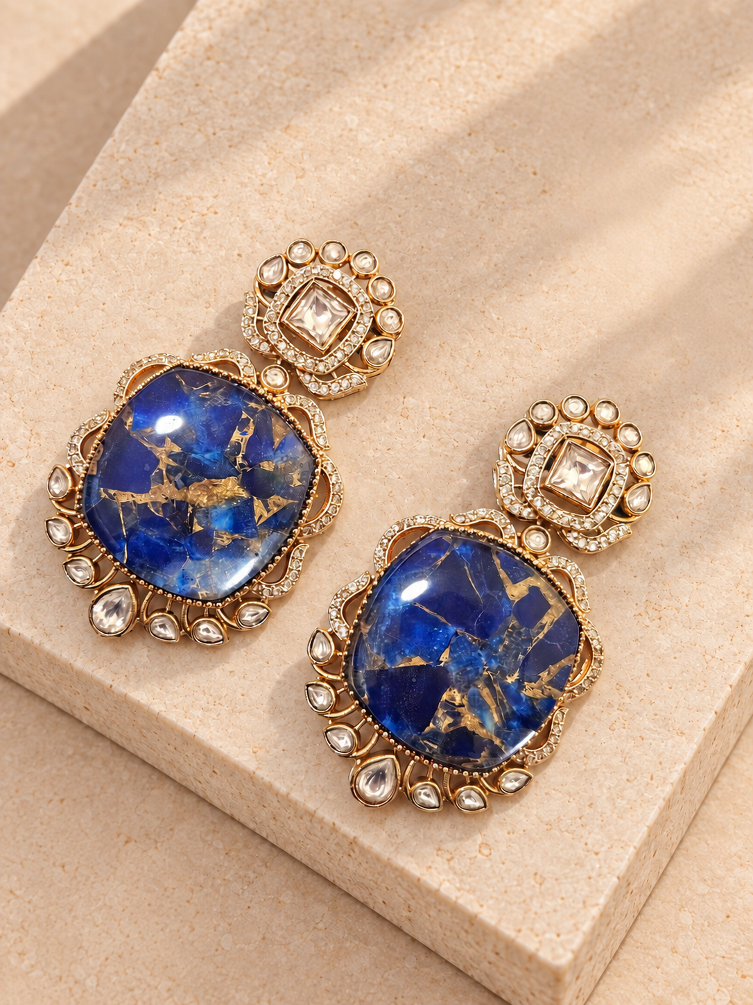 Blue doublet earrings with irregular gold-toned veining and clear stones resting on beige square stone.