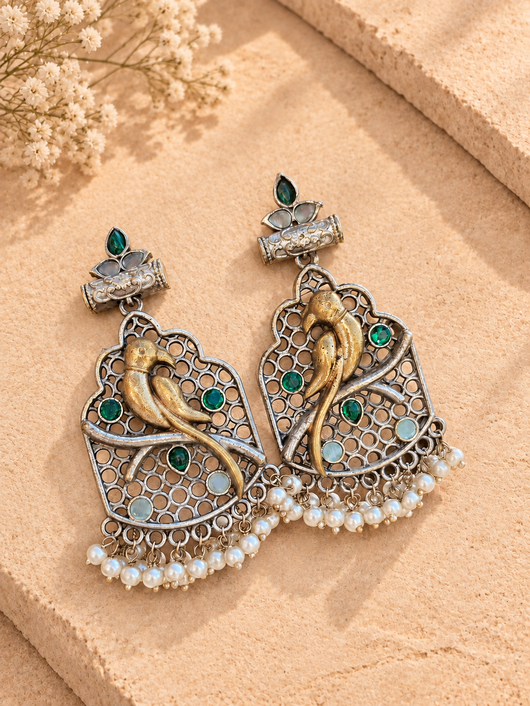 Dual-tone bird drop earrings with green and pale stones and white bead fringes, resting on beige stone.