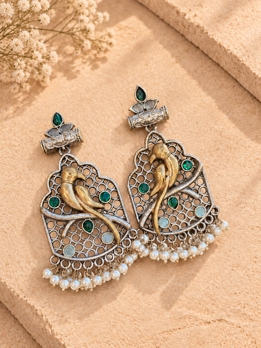 Dual-tone bird drop earrings with green and pale stones and white bead fringes, resting on beige stone.