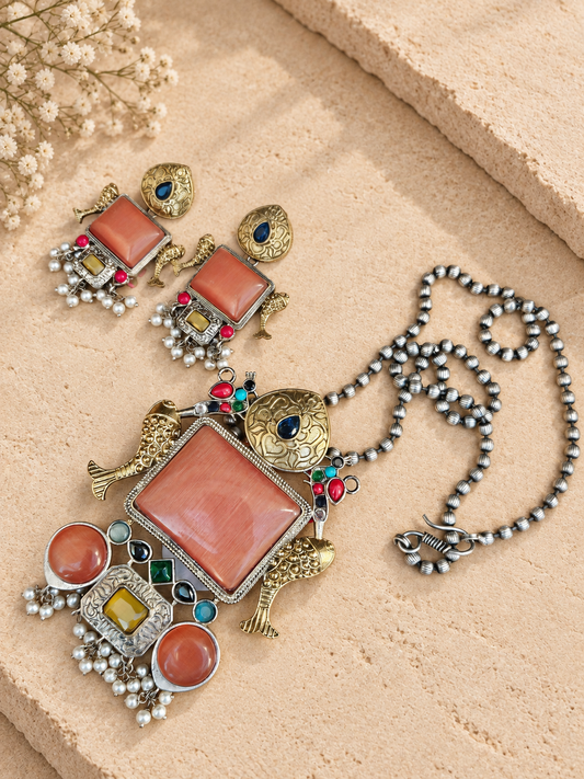 Dual-tone fish pendant set with peach-orange Monalisa stones and matching earrings on a beige stone background.